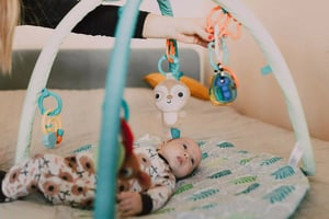 A baby lying on a patterned play mat, reaching for colourful hanging toys, including a sloth and rings, with a caregiver's hand above.