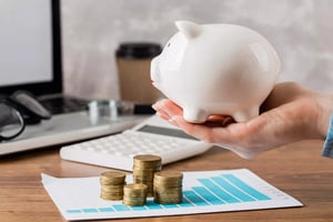 A hand holding a piggy bank next to a pile of coins