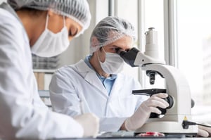 Two scientists in lab coats and masks work with a microscope, analysing samples in a modern laboratory setting.