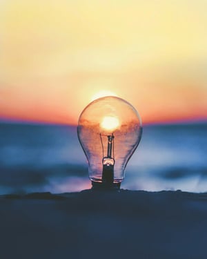 A light bulb is positioned on a surface, with the sunset reflecting inside it. The background features a blurred ocean and colourful sky at dusk.