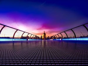 A striking, shot from the floor of the Millennium Bridge in London. The handrails are lined with glowing blue neon lights, leading the eye toward the illuminated dome of St. Paul's Cathedral in the distance. The sky above is purple, pink, and orange at twilight, with figures walking on the bridge.