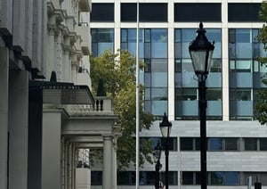 A street view featuring Falmouth Hall, a classical white building with ornate architectural details, next to a modern glass-paneled building. A black lamppost is visible in the foreground with greenery and trees in the scene.