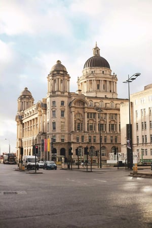 A large historic building with a prominent dome, decorative architectural elements, and ornate details, situated at the corner of a city intersection. The surrounding area includes light traffic and street signs under a cloudy sky.