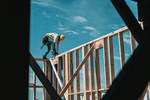 A construction worker in a yellow hard hat is positioned on the edge of a wooden frame, installing beams on a building under a blue sky with scattered clouds. The image shows wooden beams and structural elements in the foreground.