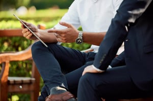 A close-up of two men seated outdoors, one wearing a white shirt and a watch while holding a tablet, gesturing with his hand. The other man in a dark suit sits beside him, legs crossed, in front of a lush green background.