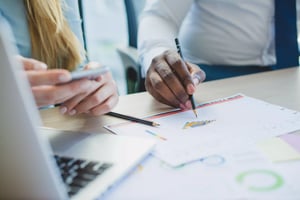 A close-up of two hands on a table during a meeting. One hand holds a smartphone, while the other is writing with a pen on a document showing colored charts. A laptop is partially visible in the foreground.