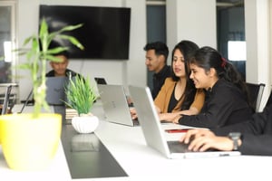 A group of professionals working at a conference table with laptops. Two women are collaborating closely, while others are focused on their screens. A green potted plant is placed on the table, enhancing the workspace environment.