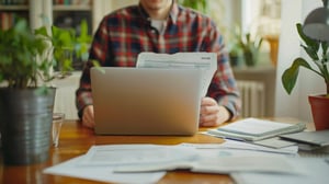 A person in a plaid shirt sits at a wooden table with a laptop open in front of them. They hold printed documents while surrounded by potted plants and scattered notebooks and papers, with natural light coming through a window.