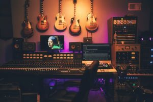 A recording studio with a large mixing console, computer screens, and audio equipment. Five electric guitars hang on the wall above, and a painting of a woman is displayed on a monitor in the center.