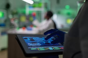 A person wearing blue gloves uses a tablet displaying a scientific image, possibly a microscopic view, in a laboratory with another person working in the blurred background.