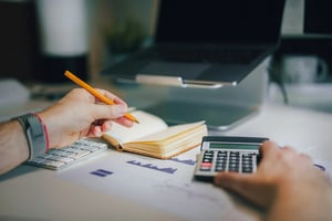 A person is writing in a notebook with a pencil while using a calculator, with a keyboard, laptop, and financial documents on the desk.