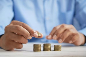A person in a blue shirt stacks coins into neat piles on a white table, suggesting concepts of saving money, budgeting, or financial planning.
