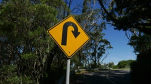 A yellow road sign with a black U-turn arrow warns drivers of a sharp curve ahead, surrounded by trees with a winding road visible in the background under a clear blue sky.