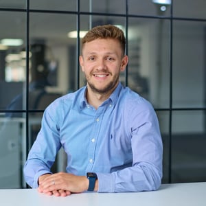 Ben Guyton, Director of Accounting in blue shirt, smiling. The background shows a grid of dark-framed glass office windows, slightly out of focus.