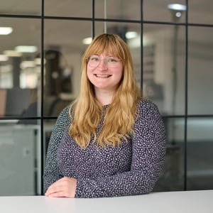 Holly Lyden, Enquiry Manager in spotted top, smiling. The background shows a grid of dark-framed glass office windows, slightly out of focus.
