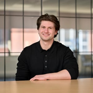 James Nash, R&D Dispute Associate in black top, smiling. The background shows a grid of dark-framed glass office windows, slightly out of focus.