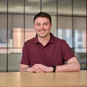 Joseph Rimmer, Client Tax Manager in red polo, smiling. The background shows a grid of dark-framed glass office windows, slightly out of focus.