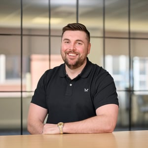 Simon Wilby, Director in black polo, smiling. The background shows a grid of dark-framed glass office windows, slightly out of focus.