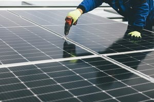 Close-up of a worker in a blue uniform and neon green safety gloves installing a grid of black solar panels. The technician is using a red and black cordless power drill to fasten a mounting bracket between two panels.